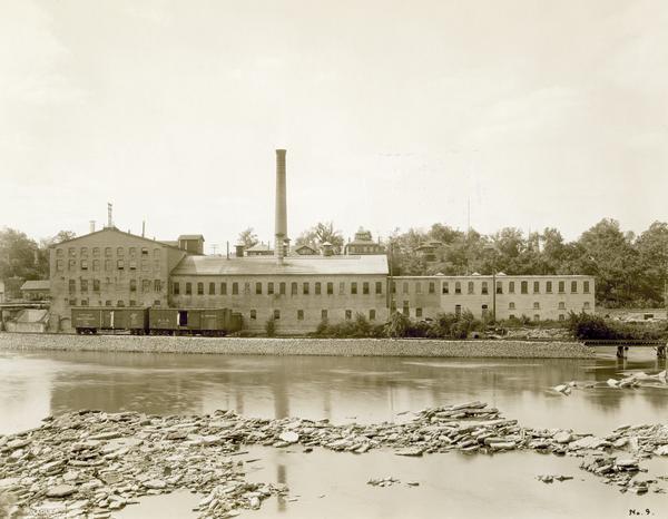 View across water towards the Atlas Mill. The paper making mill on the Fox River was built in 1878 and proved that paper could be made efficiently from groundwood pulp. Houses are on the hill in the background behind the mill.