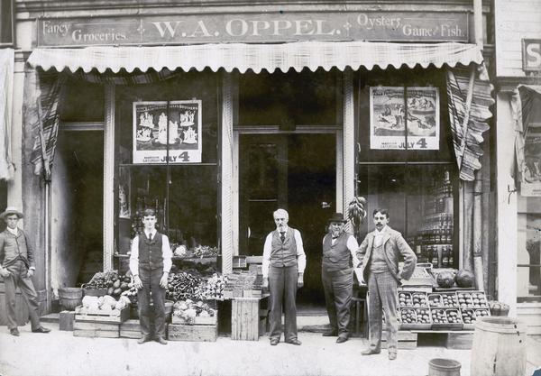 Grocers standing in front of the W.A. Oppel Grocery located at 116 East Main Street. The five men are standing on the sidewalk among bine and crates displaying vegetables and fruit. The sign above the storefront reads: "Fancy Groceries, W.A. Oppel. Oysters, Game & Fish."