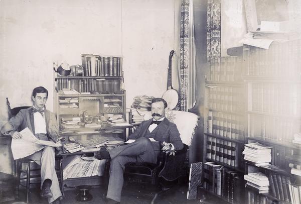 Portrait of two men sitting in a room lined with bookshelves. There is a banjo leaning in the corner next to a window.