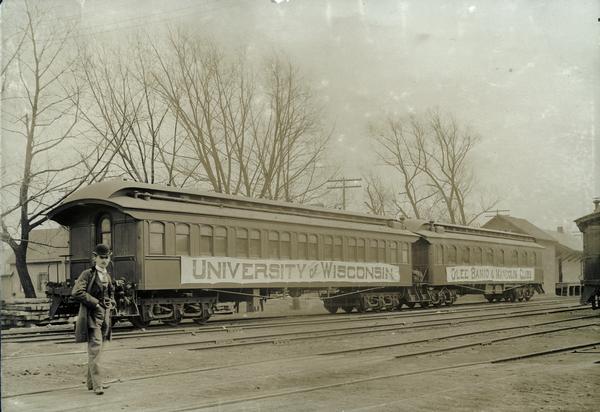 Young man posing on train tracks in front of two passenger cars decorated with banners for the University of Wisconsin Glee, Banjo, and Mandolin Club.