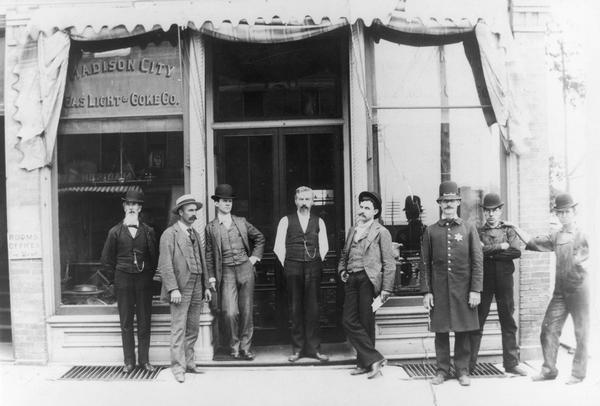 Eight men, including a police officer, pose in front of the Madison City Gas, Light and Coke Company building.