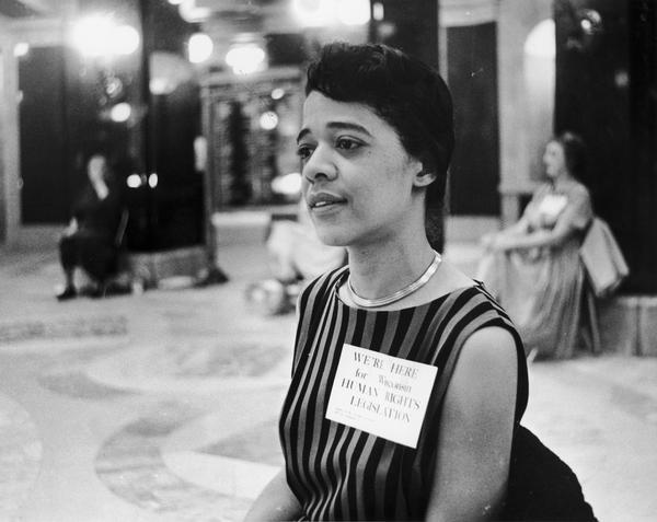 Vel Phillips wearing a striped dress and a tag that reads "We're here for Wisconsin Human Rights Legislation." Phillips was taking part in a sit-in in support of a bill prohibiting housing discrimination. The bill was defeated 53-39 on that day.