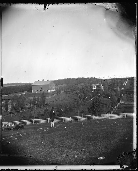 A bearded man is standing in the foreground in front of a fence, and a person is sitting on the fence on the right. Two people are standing on the hill behind the fence. A man on the far left is sitting in a horse-drawn cart on the road in front of the fence. On the hill beyond is a farmstead, including an orchard, farmhouse, and a barn with a cupola and haystacks. There are beehives among the shrubs and trees near the farmhouse. Two people are posing in the barn windows.