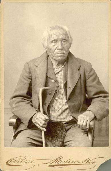 Studio portrait of prominent Winnebago man Spoon-de-Kaury, or Spoon Decorah. He is seated, holding a walking cane.