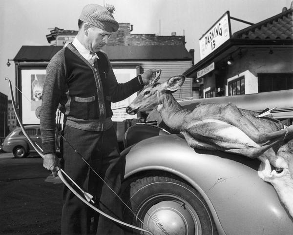 Man in a cardigan and knit hat holding a bow and arrow poses with a dead deer on the hood of a Chrysler.