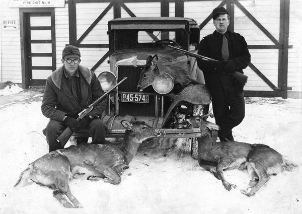 Two hunters with rifles pose in the snow by a car with three deer carcasses.