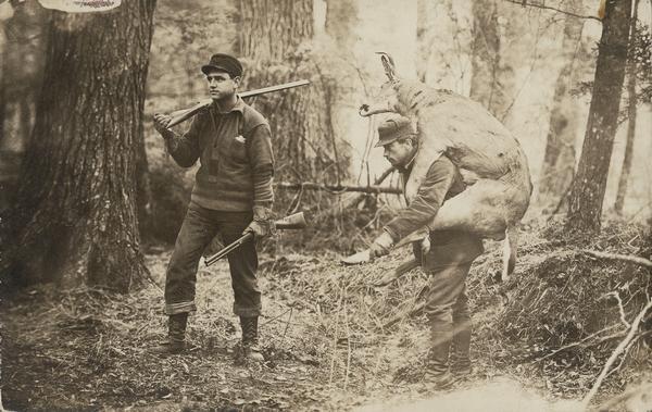 Two hunters in woods, one carrying a dead deer on his back.