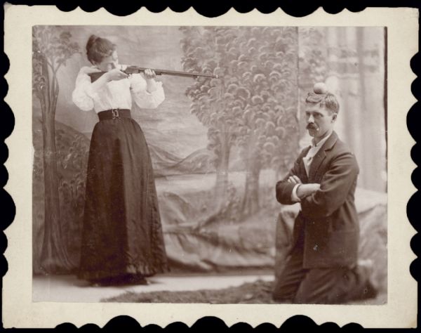 Ada [& friend] Charlie. Ada is pretending to shoot an apple from the head of her friend Charlie, who is posing kneeling with his arms folded. There is a painted backdrop behind the pair.