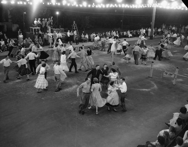 Couples folk dancing outside, with a stage in the background with musicians. Lights are strung above the dancers heads with rows of people sitting in chairs surrounding the dancers.
