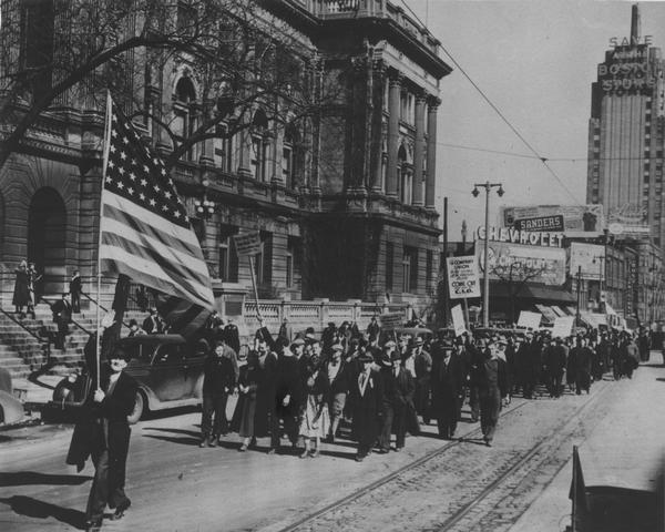 Group of men and women marching down Wisconsin Avenue. On the left is the Milwaukee Public Library, located at 814 West Wisconsin Avenue. Large sign reads: "The company union is the enemy of the American people - come out and join the C.I.O." Small sign on the left reads: "We're going places! How about you! Allis-Chalmers Workers Union" (bottom line unreadable). Small sign in the middle reads: "Organize the unorganized with the C.I.O. Allis-Chalmers Workers Union" (bottom line unreadable).