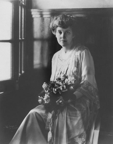 Portrait of Zona Gale, seated indoors holding flowers.