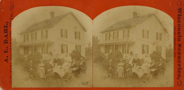 A family of eleven, including 2 babies, sits around a table set for coffee; a woman stands with coffee pot, and men hold large books and newspapers. A white, shuttered house is seen behind.