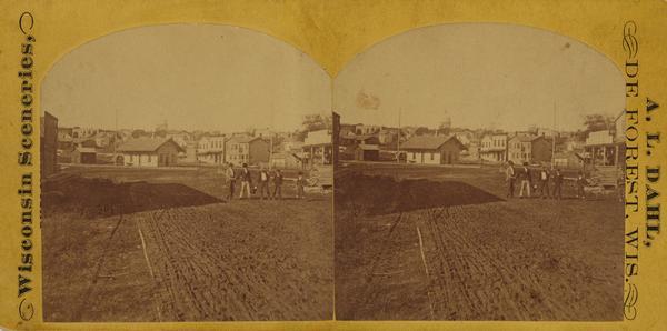 View looking southwest down the 600 block of Williamson Street to the East Madison Passenger Depot of the Chicago & Northwestern Railroad. There are frame buildings on the left and the Wisconsin State Capitol building in the background. The small brick building just under the capitol dome was the Milwaukee Road East Madison depot, built in 1869. A group of boys are posing in the road. The wood frame building on the right has a sign reading: "Charles Hinrichs Dry Goods Groceries, Boots and Shoes".