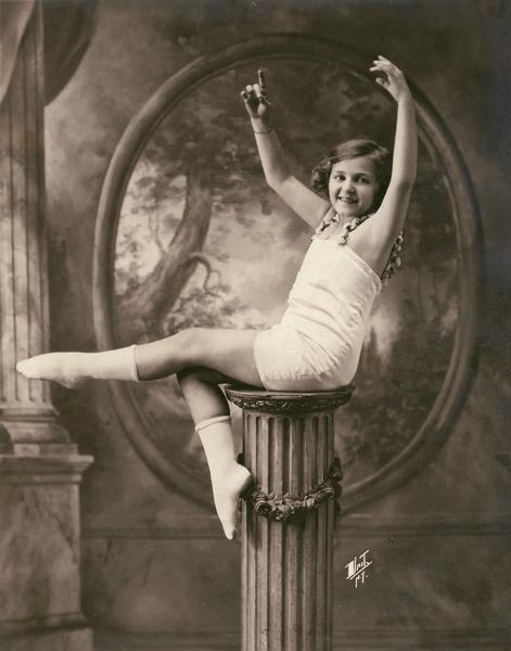 A young ballet dancer posed on a pedestal in front of a photographic backdrop. She is wearing socks over her shoes for a coordinated look. The young woman may have been a student of the Kehl School of Dance in Madison, Wisconsin.