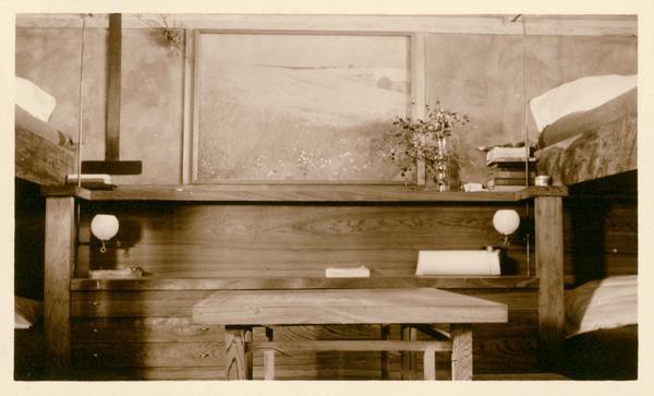 Bedroom off the end of the workroom with bunk beds on each side framing a built-in dresser stacked with books. A t-square, and a painting by George Niedecken is hanging on the back wall. A worktable in the foreground is at the center of the room. Taliesin is located in the vicinity of Spring Green, Wisconsin, and was the summer home of Frank Lloyd Wright.