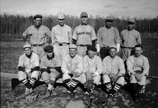 Outdoor portrait of the Wittenberg Grays baseball team, presumably taken at a baseball field.  Front row is seated on a makeshift bench.  Melvin Schlytter (catcher) is front row, far right; J. Will Gates is back row, second from left.  Back row center, wearing the White Sox jersey, is probably George "Buck" Weaver, former Chicago White Sox third baseman who was banned from professional baseball in connection with the 1919 Black Sox scandal.