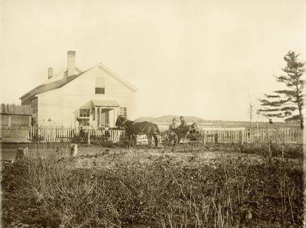 Exterior view of a farmhouse with a picket fence. In front of the house are two men and a horse-drawn carriage.