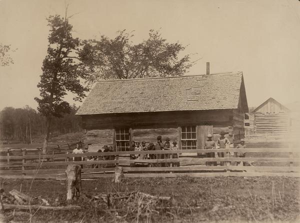 Exterior view of the last log schoolhouse in Weston with students and their teacher posing outside.