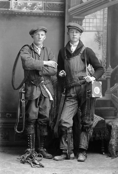 Studio portrait of two telephone linemen with some of their equipment in front of a painted backdrop. They are wearing work clothes and hats, and hold cigarettes in their hands.