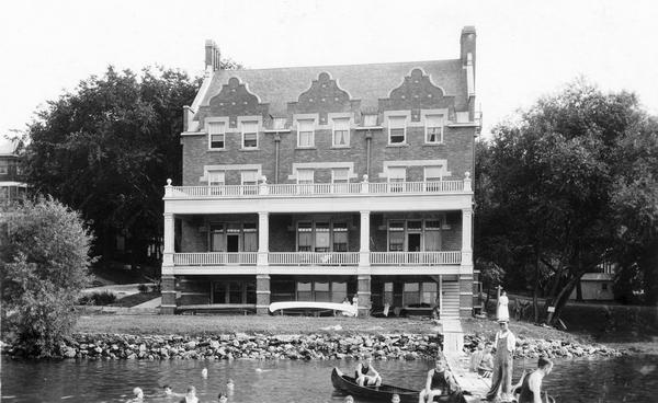 Exterior front view from Lake Mendota of the Delta Upsilon Fraternity House, located  at 640 North Francis Street, erected in 1906. A group of people are on the pier, and a man is sitting in a canoe. Other people are swimming.