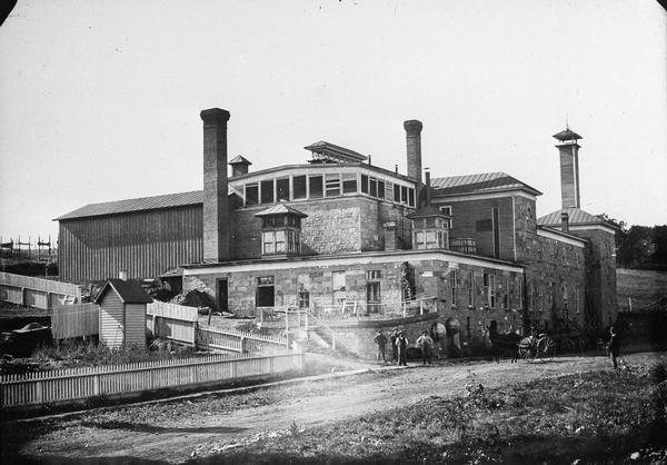 Exterior view of a brewery. Men pose in front of the building near a horse-drawn vehicle.