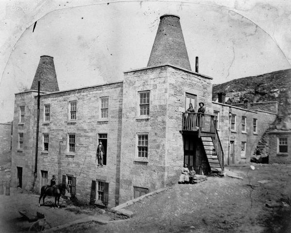 Exterior view of the Garden City Brewery, built in 1854. A person is on horseback in the yard below on the left, and a man is standing above them in an open second-story doorway. Other people are posed at the top of the steps in the center, with two people sitting on the stone wall below.