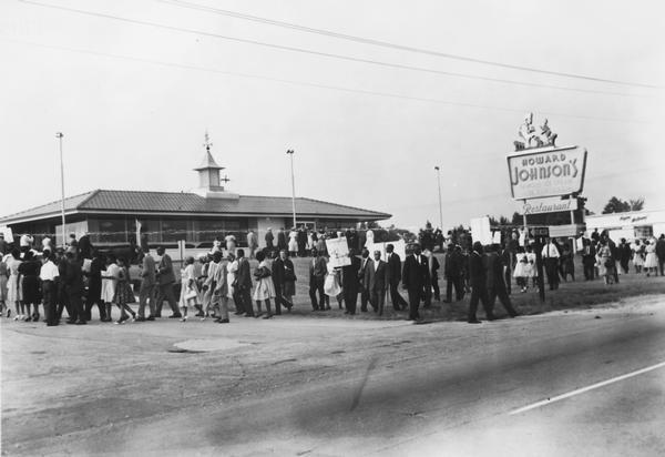 Group of protesters march in front of a Howard Johnson's restaurant holding signs. One says "Segregation OUT!! Integration IN!! CORE - NAACP".