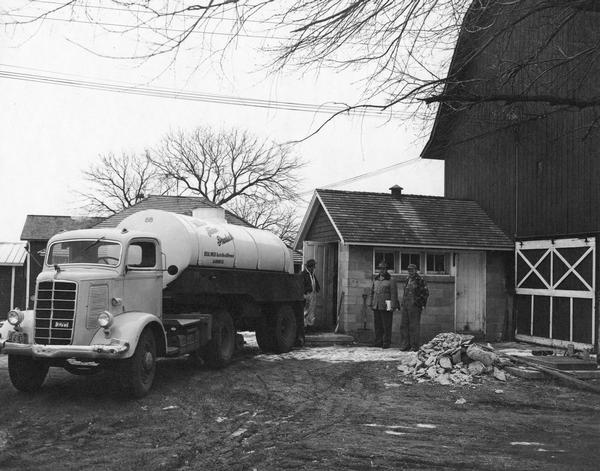 A bulk milk truck is parked in a farm yard near a barn as three men stand by.