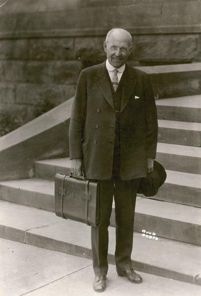 Professor Stephen Gilman stands on steps holding his hat and briefcase.