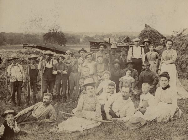 A large family group and workers pose in field in front of farming machinery and haystacks.