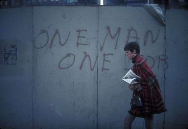A business woman clutching her purse and a newspaper walks past a large portion of concrete with "One Man, One Job" painted on it. This is at the site of the World Trade Center construction.
