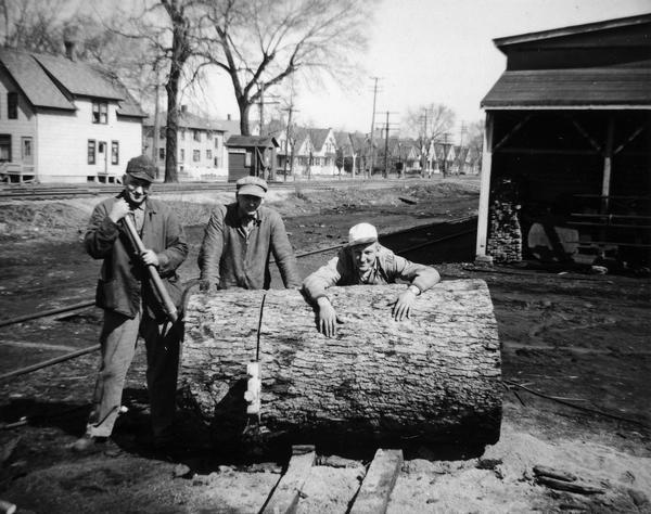 Joe Hess, Eddie Hess, and Foots Hess (l to r) of the Hess Cooperage pose with a large oak log which will be cut into staves and heads for barrels. Houses in the background are along the 1900 block of East Main Street.