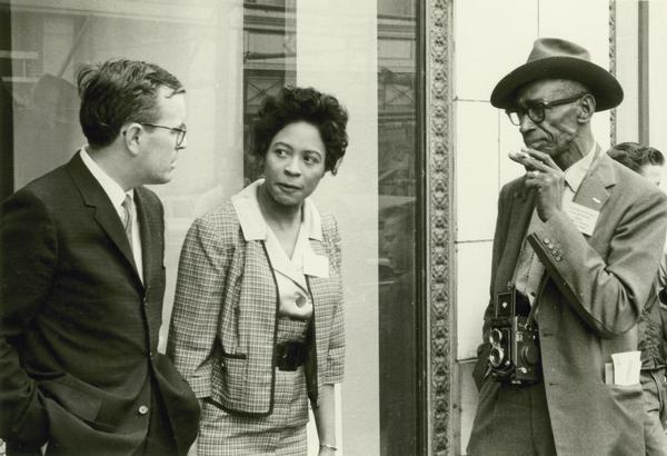 Daisy Bates and L.C. Bates, apparently wearing press badges, standing outdoors talking with an unidentified man. L.C. Bates, who is wearing a camera, is apparently present as a photographer for the "Arkansas State Press," owned and operated by the Bates.