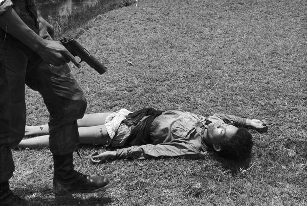 A dead Vietnamese man lays on the ground as a paratrooper prepares to fire bullets into the body.