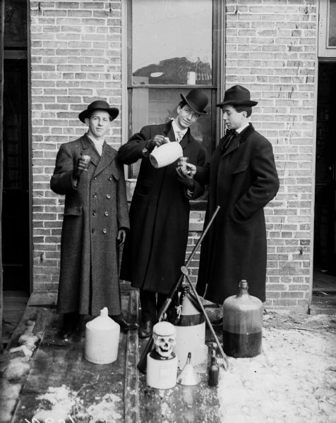 Three men in long coats and hats are standing outdoors and staging an elaborate temperance portrait, which includes jugs and an artificial human skull.
