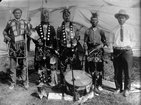 Five Ho-Chunk performers in front of cloth barriers or tents. A powwow group from the 1908 Homecoming. The five are full brothers according to the 1881 tribal rolls. Men are from left to right: Jim Swallow, William Massey(Massie), Thomas Thunder, George Eagle and Ben Thunder Cloud.  They are all dressed in regalia, except for Ben Thundercloud, who is wearing a shirt, tie, and hat.