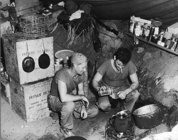 Two men make coffee in a ramshackle hospital kitchen, which consisted of a single, improvised burner. The grill is the innards of a human blood shipping case. These cases also served as storage space.
