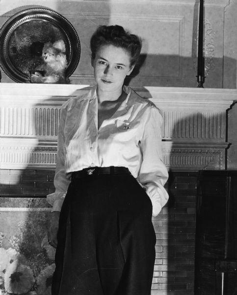 Three-quarter length portrait of a young Dickey Meyer Chapelle posing in front of a fireplace mantel wearing a blouse and dress pants.