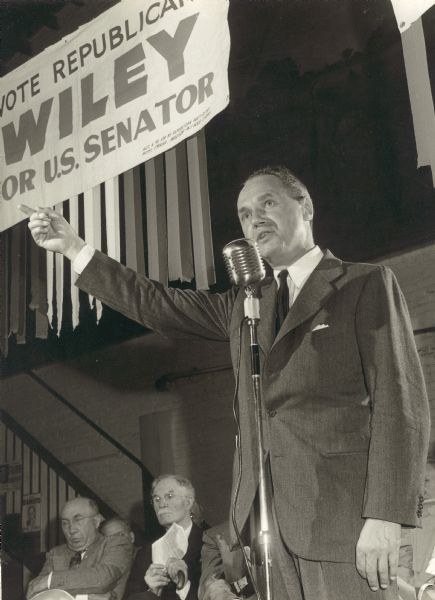 Walter J. Kohler, Jr., campaigning during the 1950 Republican gubernatorial primary at the Sales Pavilion in Waukesha.  Kohler is speaking into a microphone with his right arm raised to make a point.  Seated on the platform behind him are Secretary of State Fred R. Zimmerman and State Senator Chester E. Dempsey of Waukesha.