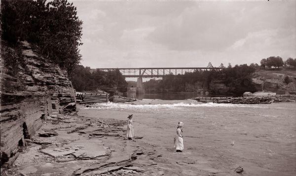 Below Kilbourn log dam, 1890s. Two girls are standing on the edge of the river. There is a bridge in the background.