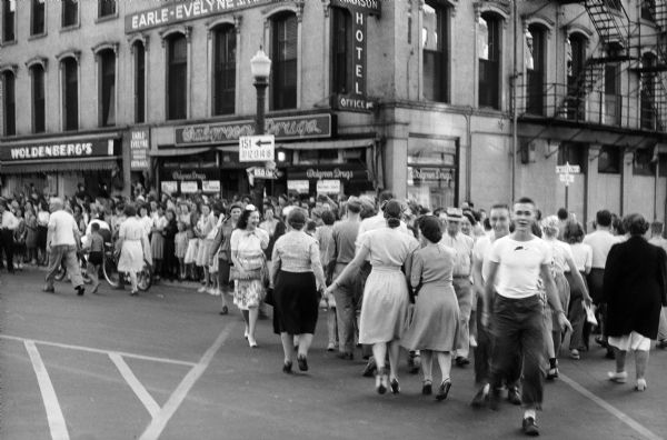Crowds at the corner of East Mifflin Street and North Pinckney Street on V-J Day, August 15, 1945, the day on which the Allies announced the surrender of Japanese forces during World War II. The building on the corner is the Madison Hotel, with the following businesses on the ground floor on Mifflin Street: Woldenberg's and Walgreen Drugs. In the upstairs window is a sign for Earle-Evelyne Salon, complete beauty care.