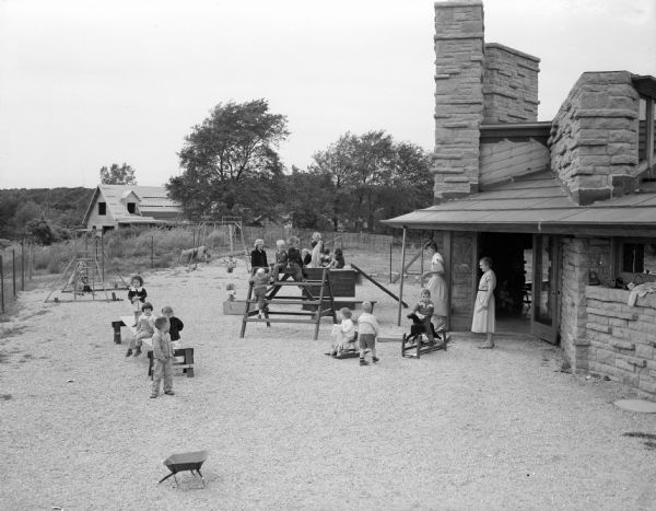 Children playing outside the nursery school classroom at the First Unitarian Society meeting house while teachers are watching. The building was designed by Frank Lloyd Wright.