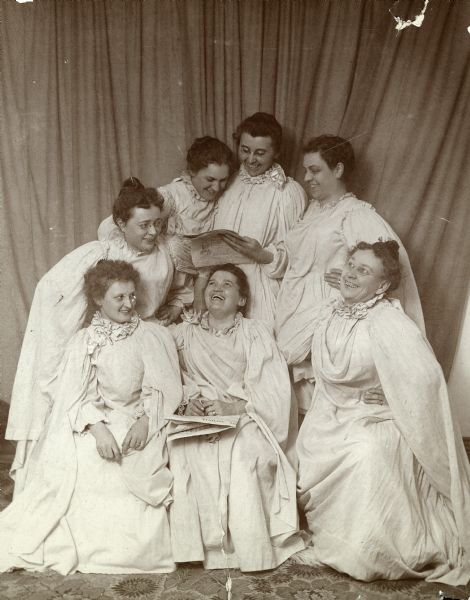 A group of seven women dressed in white flowing gowns strike "Grecian poses".