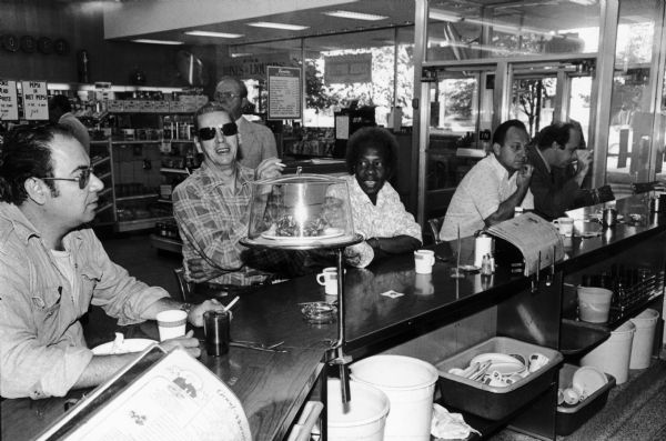 People drinking coffee and smoking at the lunch counter of the Rennebohm Drug Store #2, 30 West Mifflin Street, then known as the 30 on the Square Building. The camera angle affords a view of the rear of the counter and the drug store behind them.