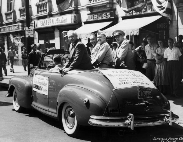 Governor Oscar Rennebohm rides in a car with several military service men in a parade.