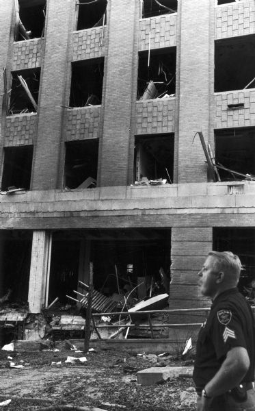 A policeman stands in front of Sterling Hall on the University of Wisconsin campus.  Early on August 24th, 1970, a van loaded with six barrels of explosives blew up just outside the East Wing of Sterling Hall at the University of Wisconsin campus in Madison. The bombing was carried out by four men in protest of America's involvement in the Vietnam War. The four men responsible were, Karl Armstrong, Dwight Armstrong, David Fine and Leo Burt.  Unfortunately, despite an attempt to detonate the bomb when the building was vacant, a physics researcher conducting research unrelated to Army Math Research Center, was killed in the explosion. The sobering impact of Robert Fassnacht's death brought a sudden halt to the violence to which anti-war protesters and police had resorted.  The bombing was directed against the Mathematics Research Center, a U.S.-Army-funded facility, which was located in the East Wing of Sterling Hall along with the physics and astronomy departments. "Army Math," as it was known, was despised by many antiwar activists who felt the center was contributing to the death and destruction in Southeast Asia through its research and had no place on a public university campus.
