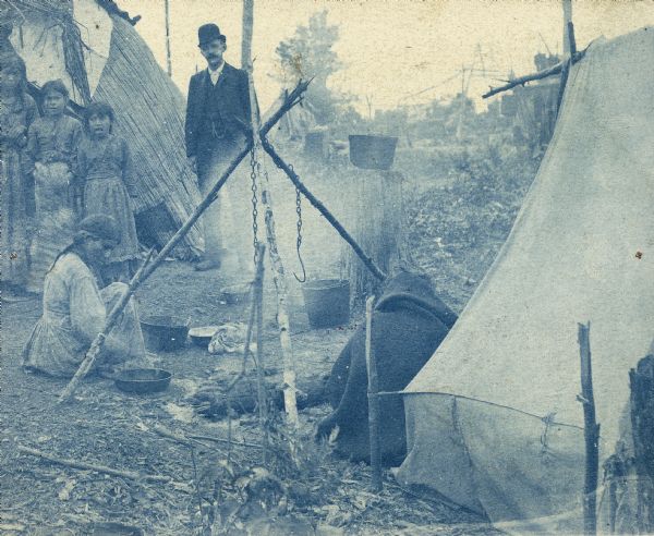 Cyanotype print of Chippewa (Ojibwa) Indians near Bradley Junction, near Minocqua. A man wearing a suit and hat is standing in the background.