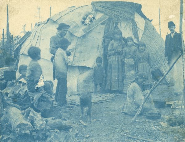Cyanotype print of Chippewa (Ojibwa) Indians near Bradley Junction which is near Minocqua. The group is standing in front of a wigwam, and a woman in the right foreground is sitting near a cooking fire. On the far right is a man standing and wearing a suit and hat.