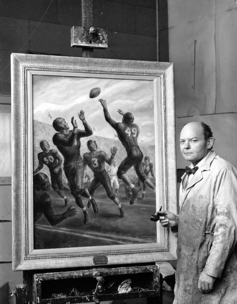 John Steuart Curry standing next to an action painting of David N. Schreiner, an All-American end in 1942 who was killed in action on Okinawa in 1945, while serving as a lieutenant with the 6th Marines. Schreiner is depicted catching a forward pass near the goal line during a football game at Camp Randall. It was originally painted for Abbott Laboratories for use in a magazine they published.  Mr. Curry suggested that they present the painting to the University of Wisconsin-Madison athletic department. Schreiner's No. 80 was retired in 1945 and again in 2006 where it adorns the Camp Randall facade with 5 other football heroes.