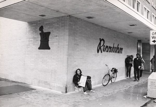 On a rainy day in 1970 a young man hawking newspapers takes cover under the overhang of the Rennebohm Drug Store at 676 State Street.  After having being damaged by anti-war violence on several occasions during the 1960s, Rennebohm's covered their glass windows with a brick facade.